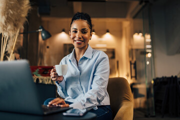 Obraz premium Portrait of a smiling African businesswoman. holding a cup of coffee, using the laptop.