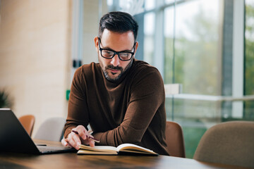 Focused businessman reading notes in a notepad, sitting in front of the laptop indoors.