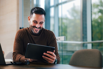 Smiling male employer using a digital tablet, taking a work break, sitting at the office.