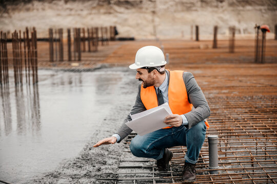 An architect is crouching next to a fresh concrete building foundation and checking on it while holding paperwork.
