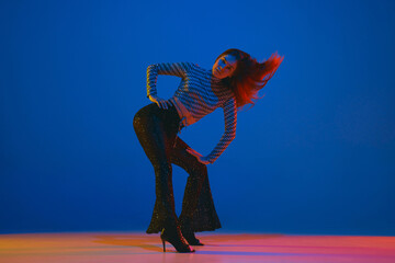 Dynamics. Portrait of young girl dancing high heel dance in stylish clothes over blue background in neon light. Concept of modern dance style