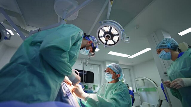 Main neurosurgeon sits at the patient while assisting doctor stands beside. Nurse in mask passing the tool to the doctors. Low angle view.