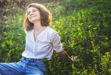 Happy pretty young cheerful woman in white shirt sitting relaxing on grass enjoying sunny spring day
