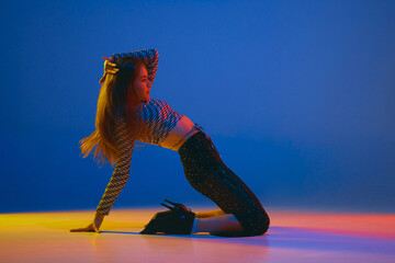 Portrait of young girl dancing high heel dance in stylish clothes over blue background in neon light. Concept of modern dance style