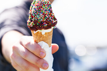 Person holding out soft serve ice-cream in waffle cone with chocolate and sprinkles