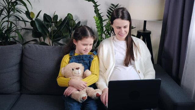 Happy pregnant mother and cute little daughter using pc laptop computer having video call conversation to family. Beautiful smiling young mom with adorable girl child sitting on cozy couch at home