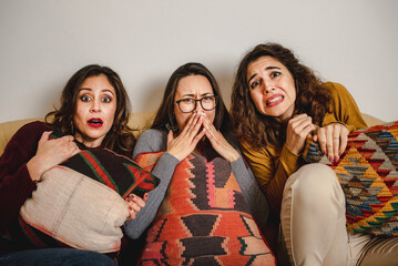 Group of frightened female friends sitting on sofa at home and watching horror movie together while...