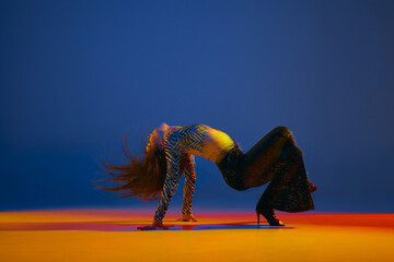 Flexibility and passion. Young girl dancing heels dance in stylish clothes over blue background in neon with mixed lights. Concept of modern dance style