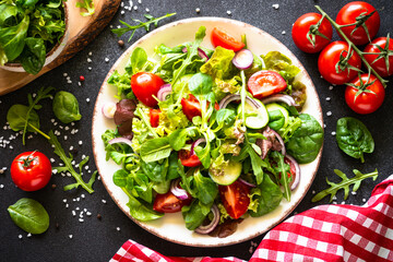 Salad leaves and vegetables in white plate at black table. Top view with ingredients.
