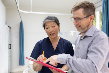 middle aged woman wearing blue scrubs holding a file while showing it  to a man in a clinic ward