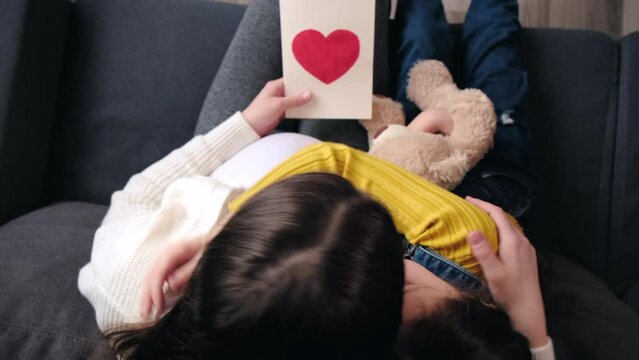 Top view of little girl hugging young mother, congratulating, presenting greeting card with wishes, mum and loving daughter sitting on cozy sofa. Family celebrating birthday or women mothers day