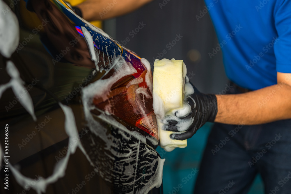 Wall mural Man worker washing car service with foam and sponge. Car wash cleaning wipe station. Employees clean a vehicle professionally.