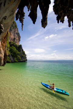 Bikini clad female cuts through the tropical water in a kayak. Stalactites hang overhead as she kayaks around the rock.