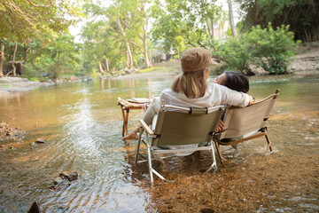 Pleased happy mother and daughter sit camp chair looking out over river, Soak your feet in the river, sit near a camp and a tent, drink coffee in a pine forest. Camping, recreation, hiking.