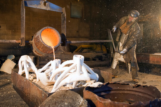 A man pouring orange molten bronze into white moldings in a foundry with the help of a support crane.