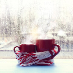 small and large red mugs are wrapped in a scarf against the background of a window with raindrops. together in the warm morning atmosphere