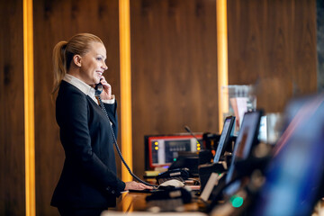 A happy receptionist is talking on the phone with hotel guest while standing at reception.