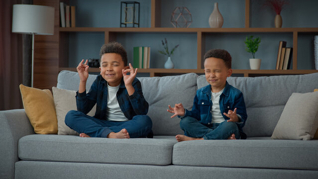 Two Funny Little African American Ethnic Children Boys Brothers Siblings Kids Meditating Together Sitting On Couch In Lotus Position. Small Schoolboys Meditate. Spiritual Development Yoga Meditation