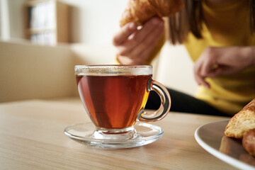 A cup of tea on a table indoors, with teenage girl eating croissant