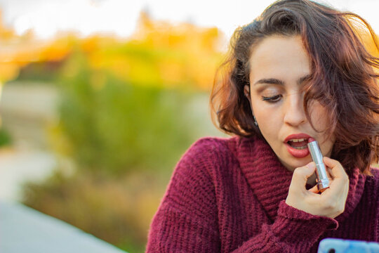 Mujer joven de raza cauc&aacute;sica con el pelo corto pint&aacute;ndose los labios con un pintalabios rojo en un parque de Madrid mientras espera a su cita de San Valent&iacute;n.