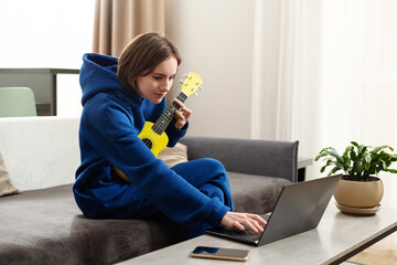 A teenager girl is engaged in her favorite hobby at home, learning to play the ukulele or guitar remotely using video on the Internet. She plays music, takes new skills, recreation