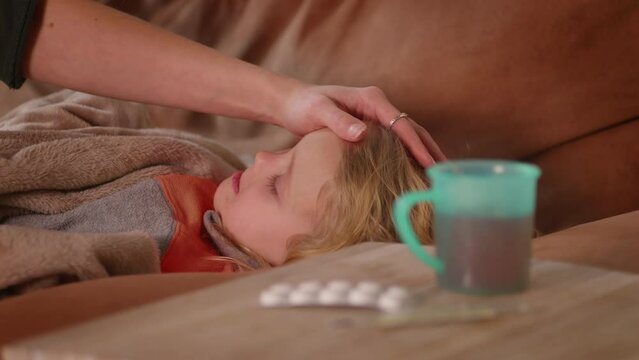 Close up view of sick child lying on the sofa with fever, mother touches her head with a hand, caring tender family moment, hot drink, pills and thermometer. Corona symptoms, virus disease. HQ 4K
