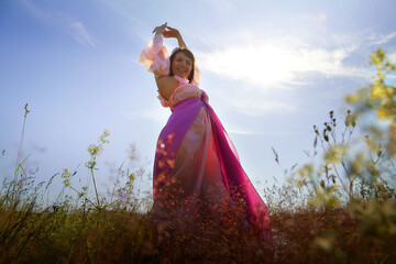 Beautiful girl in a lush pink ball gown in green field during blooming of flowers and blue sky on...