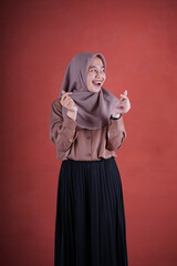 Fototapeta na wymiar Beautiful Asian woman in brown shirt and hijab smiling cheerfully showing Korean heart with two fingers crossed, express joy and positivity on brown background