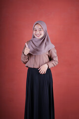 Beautiful Asian woman in brown shirt and hijab smiling cheerfully shows upset expression with thumbs up on brown background