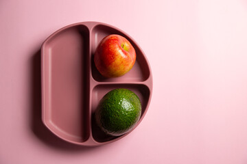 The top view of modern silicon eco baby dish with avocado and apple on the pink background. Self feeding concept, copy space, vertical view - obrazy, fototapety, plakaty