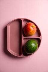 The top view of modern silicon eco baby dish with avocado and apple on the pink background. Self feeding concept, copy space, vertical view