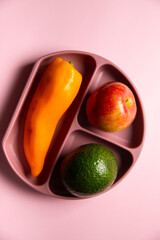 The top view of modern silicon eco baby dish with pepper, avocado and apple on the pink background. Self feeding concept, diagonal view