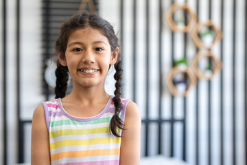 Portrait of cheerful girl with pigtails hair in colorful dress look at camera in room. Smiling kid relaxing at home.