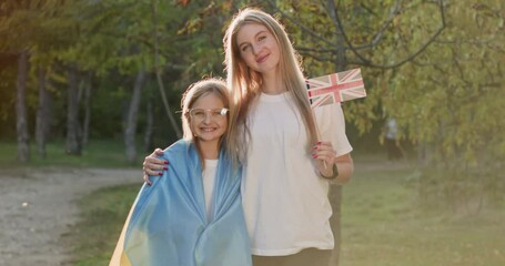 Two girls are hugging, an adult girl is hugging a little girl, a girl with glasses is wrapped in a fabric flag. An adult girl holds the flag of the United Kingdom in her hand.