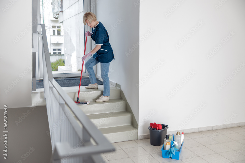 Wall mural cleaning lady in uniform with mop and rag washes a dusty steps of the house