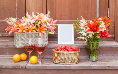 Lilies, astilbe, strawberry, oranges, white frame and rose wine on steps of rustic wooden ladder. View with copy space.