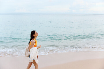 Fototapeta na wymiar Woman smile with teeth after swimming in the ocean with a backpack in a wet yellow tank top and denim shorts walks along the beach, summer vacation on an island by the ocean in Bali sunset