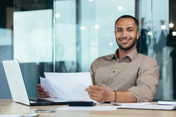 Portrait of young successful hispanic businessman inside office, man smiling and looking at camera, paper worker happy with achievement results sitting at workplace with laptop.
