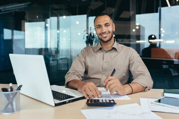 Fototapeta na wymiar Portrait of young successful hispanic businessman inside office, man smiling and looking at camera, paper worker happy with achievement results sitting at workplace with laptop.