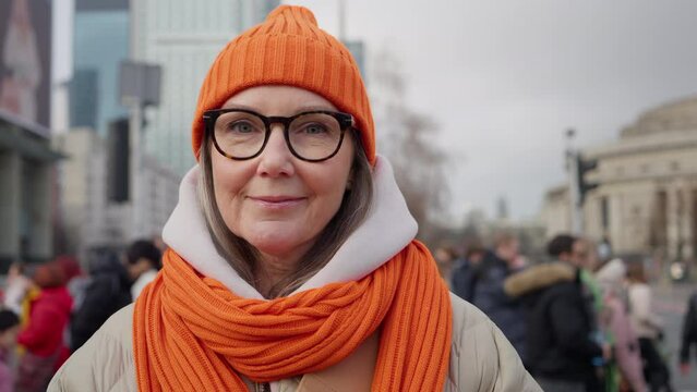 Portrait Urban Style Happy Elderly 60s Female Model Standing On Busy City Street. Senior Woman In Trendy Winter Outfit Orange Hat Scarf Backdrop Blurred People Crowd Crossing Crosswalk Slow Motion