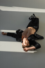 high angle view of man in black pants and blazer on shirtless body lying in sunlight on grey background.
