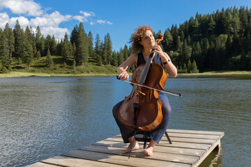 Young beautiful woman playing the cello outdoors in the mountains on a wooden jetty with a lake and beautiful nature behind it. © alexandre zveiger