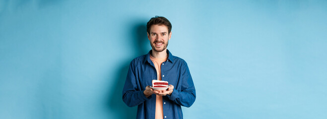 Celebration and holiday concept. Smiling man holding birthday cake with candle and looking happy, standing on blue background - Powered by Adobe