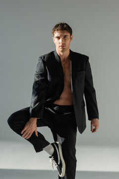 sexy brunette man in black pants and blazer on shirtless body standing on one leg and looking at camera on grey background.
