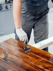 Man paints wooden boards with paint brush. Carpenter cabinetmaker varnishes wooden surface. Authentic workflow..