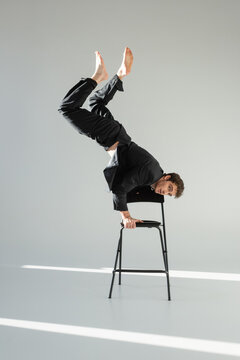full length of barefoot man in black suit doing handstand on chair and looking at camera on grey background.