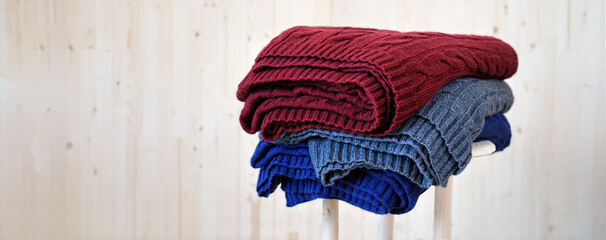 Stack of warm, multicolored pullover knitwear, pile of handmade wool sweaters on stool with light wooden background