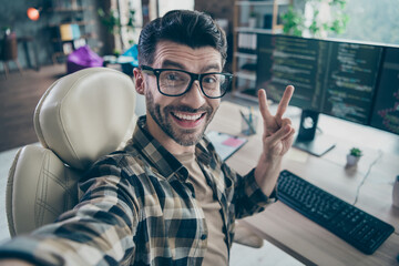 Photo of cheerful web designer guy sit chair take selfie demonstrate v-sign open space workstation indoors