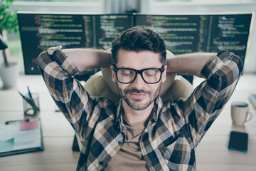 Photo of happy smiling freelancer wear eyeglasses arms hands behind head having rest after working modern gadget indoors workplace workstation loft