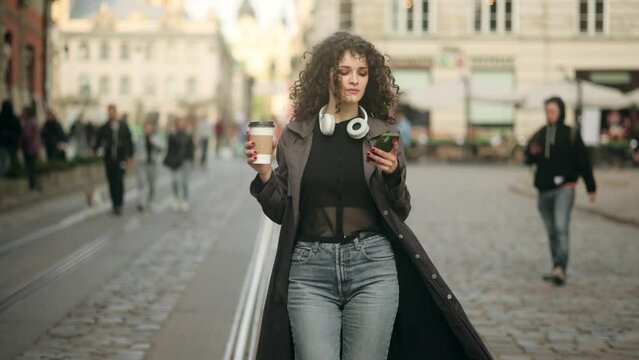 Beautiful cheerful young woman walking in the city, holding takeaway coffee cup. Joyful caucasian woman smiling and drinking coffee takeaway while walking at city street